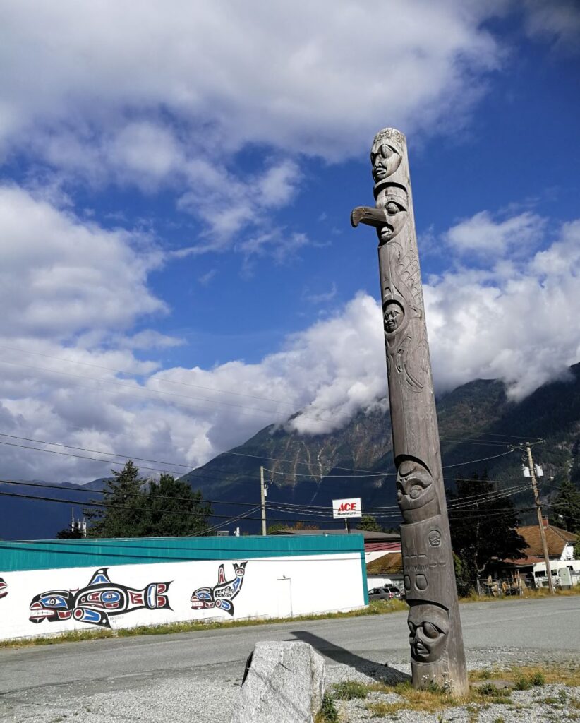 The story pole of Nuxalk People who remember the history of residential school and recognize their own strength and resilience. The pole has a mother at the bottom, 4 children above her, a father upside down without mouth, blank space in the middle, ravens, creator and four carpenters atop of the pole